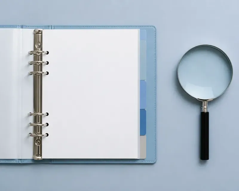 An open ring binder with tabbed dividers beside a magnifying glass